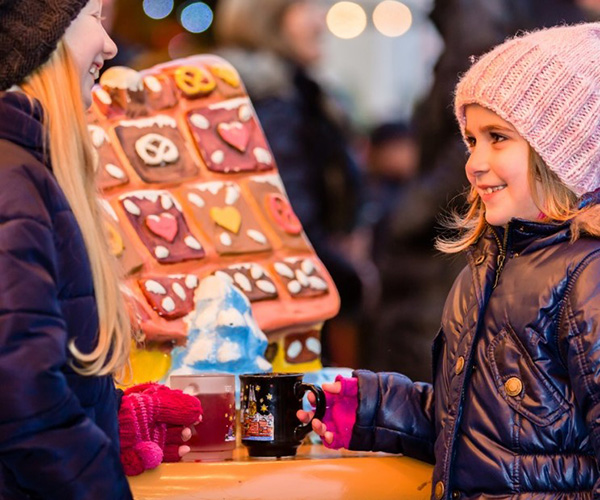 Colour image - Two young girls in winter clothing in front of a festive gingerbread house with mugs of cocoa